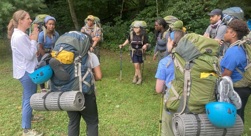 A group of people wearing backpacks stand in a circle and listen to a person give instructions. 